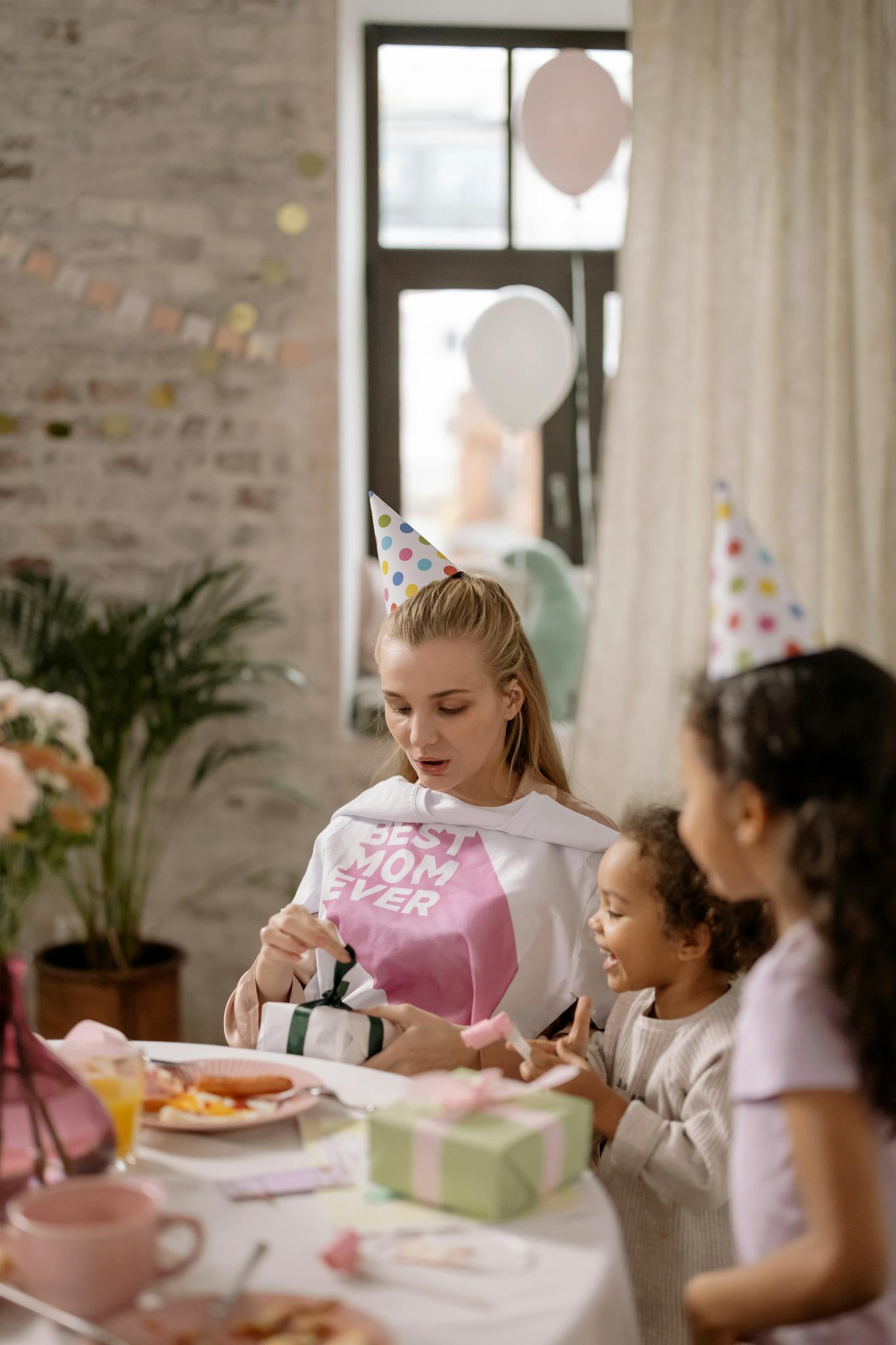 A joyful birthday party scene with a mother and daughters opening gifts indoors.