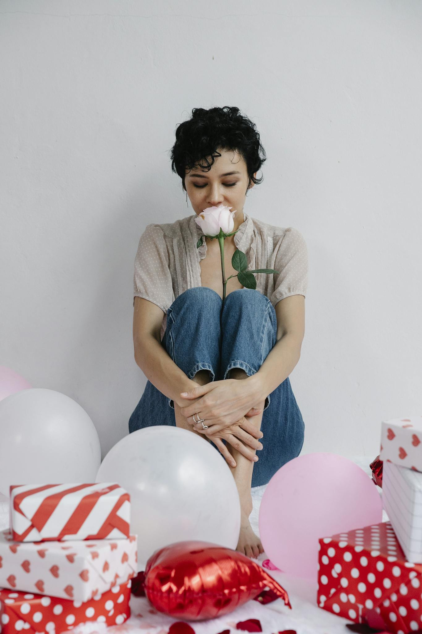 A woman sits surrounded by gifts and balloons, savoring a pink rose on Valentine's Day.